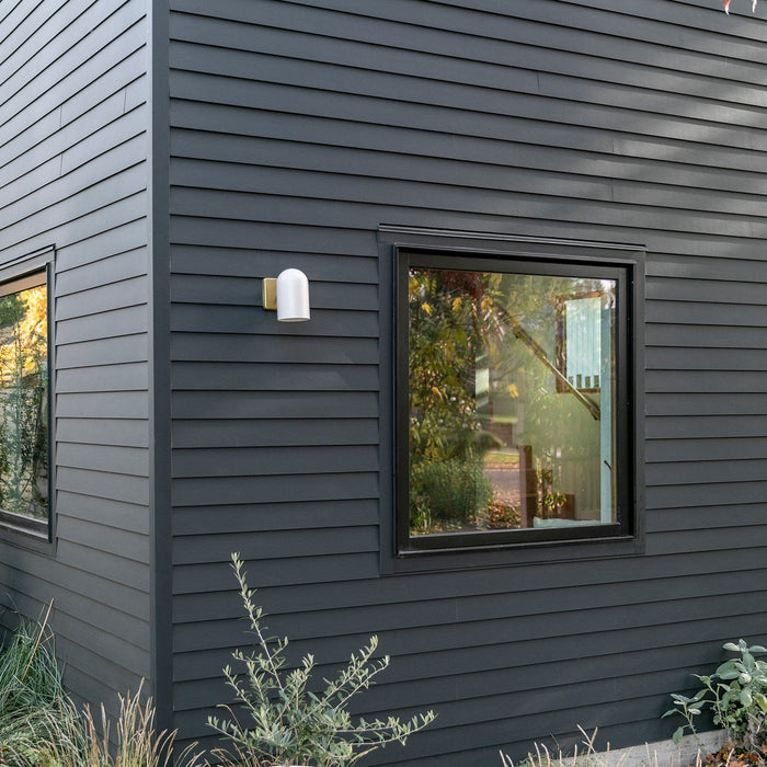 White outdoor wall sconce with brass backplate mounted on dark gray horizontal siding next to a window