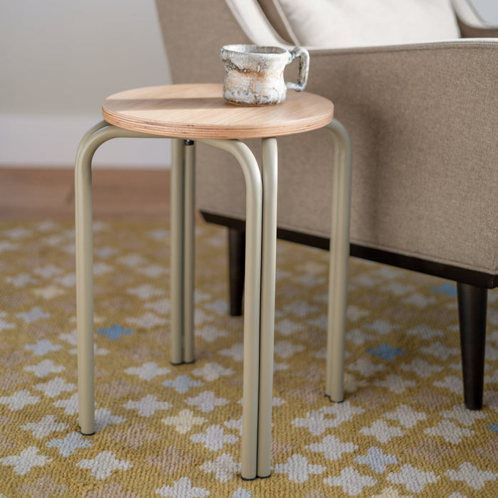 Close-up of Forma Stool showcasing white oak round top with exposed plywood edge and powder-coated steel legs beside an upholstered chair