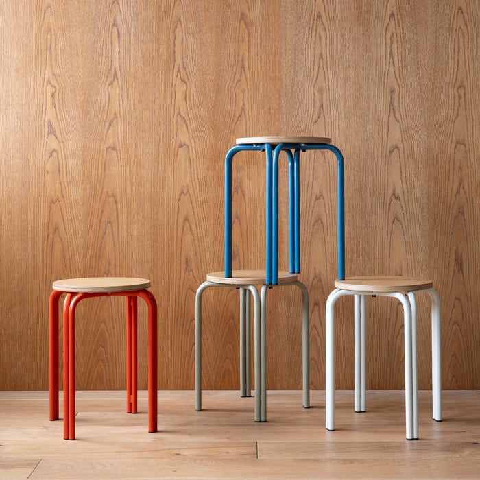 Three-quarter view of Forma Stool showing oak top and powder-coated steel legs in assorted colors against wood-paneled wall