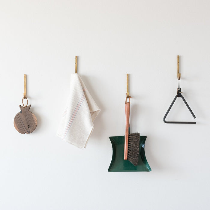 Four natural brass J hooks mounted on a white wall holding a wooden cutting board, a white tea towel, a brush and green dustpan, and a black metal triangle