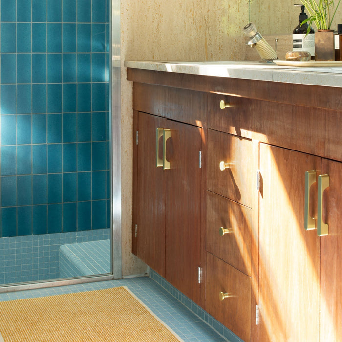 Angled view of Roland cabinet knobs in natural brass on a dark wood bathroom vanity with blue tile