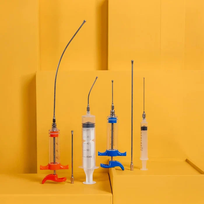 Image of a straight and curved feeding needles in a variety of sizes attached to different types of syringes, all against a bright yellow-orange background.